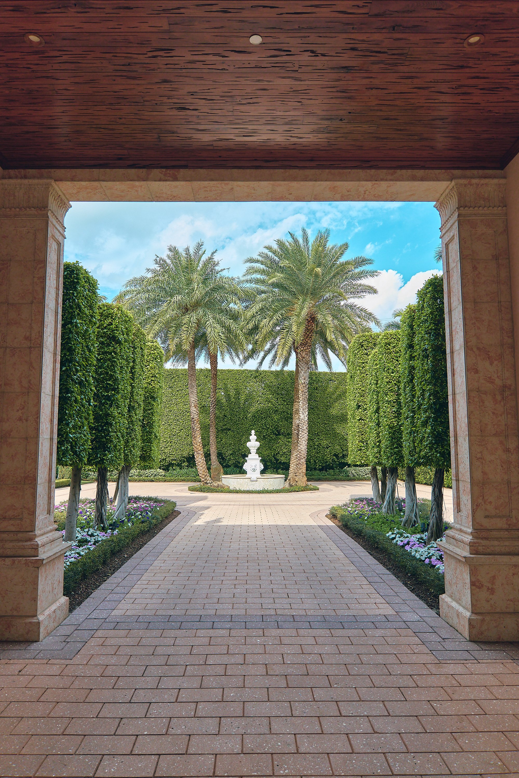 A tiled walkway leading to a court yard with palm trees and a white fountain, framed by stone columns