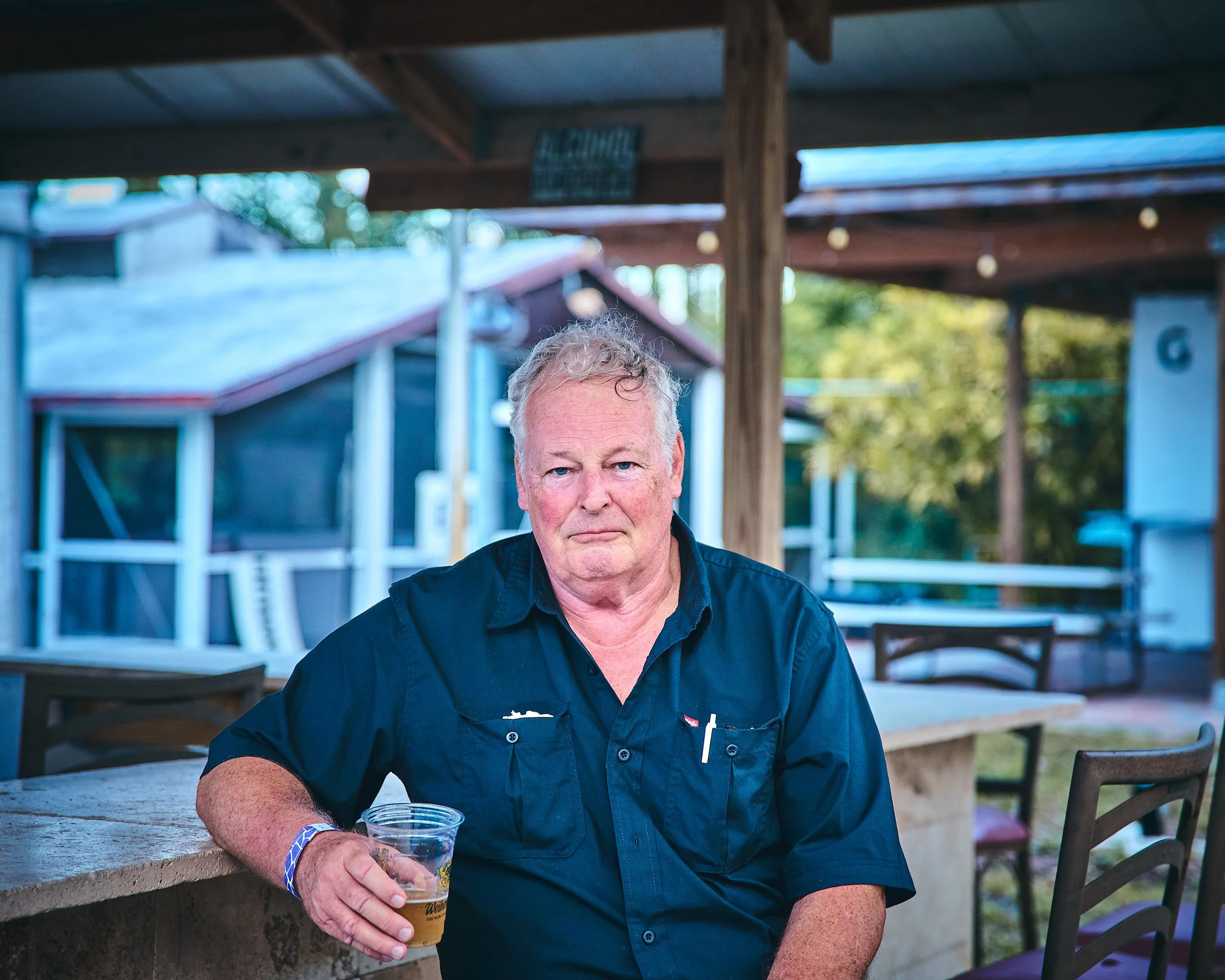 An older man with gray hair wearing a black shirt sitting at an outdoor bar or patio holding a glass of beer.