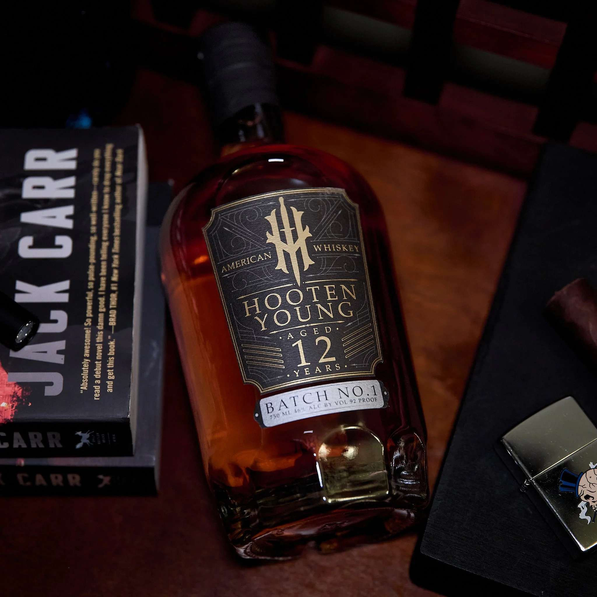 A bottle of Hooten Young whiskey aged 12 years on a wooden surface. To the left, there is a book titled 'Jack Carr' and to the bottom, part of a metallic object with a cartoon character on it.