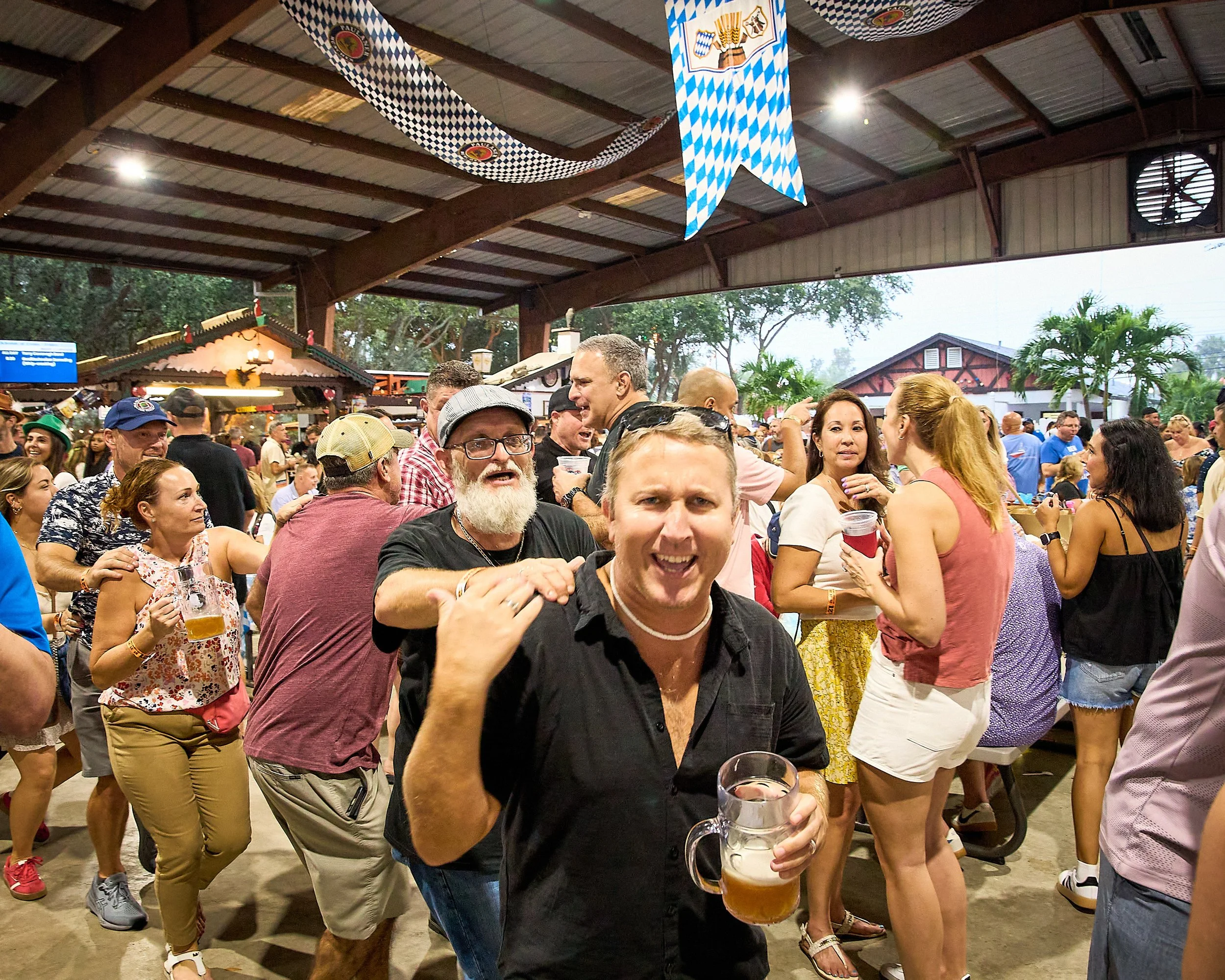 Crowd of people celebrating at an indoor festival, some holding beer mugs, with Bavarian flags and decorations hanging from the ceiling.