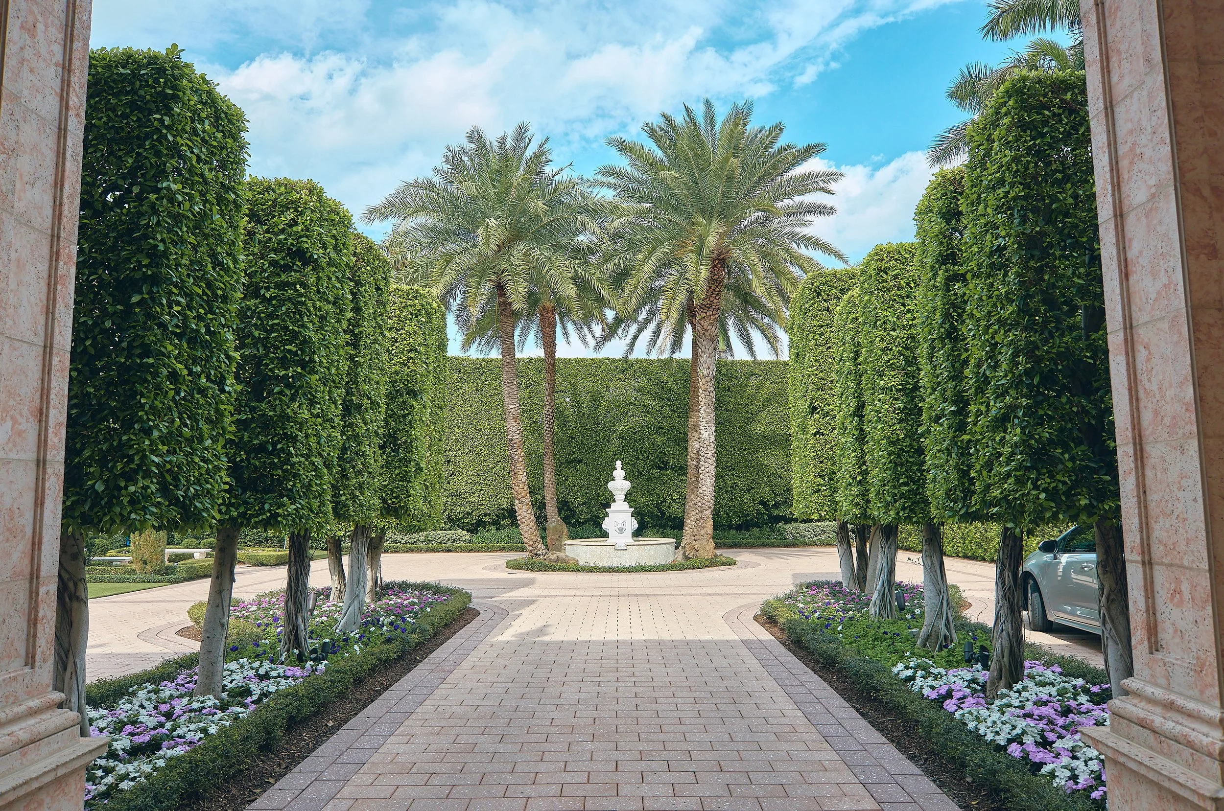 A landscaped garden with a paved pathway, green trees, and tall palm trees, viewed through a stone archway with a water fountain in the background.