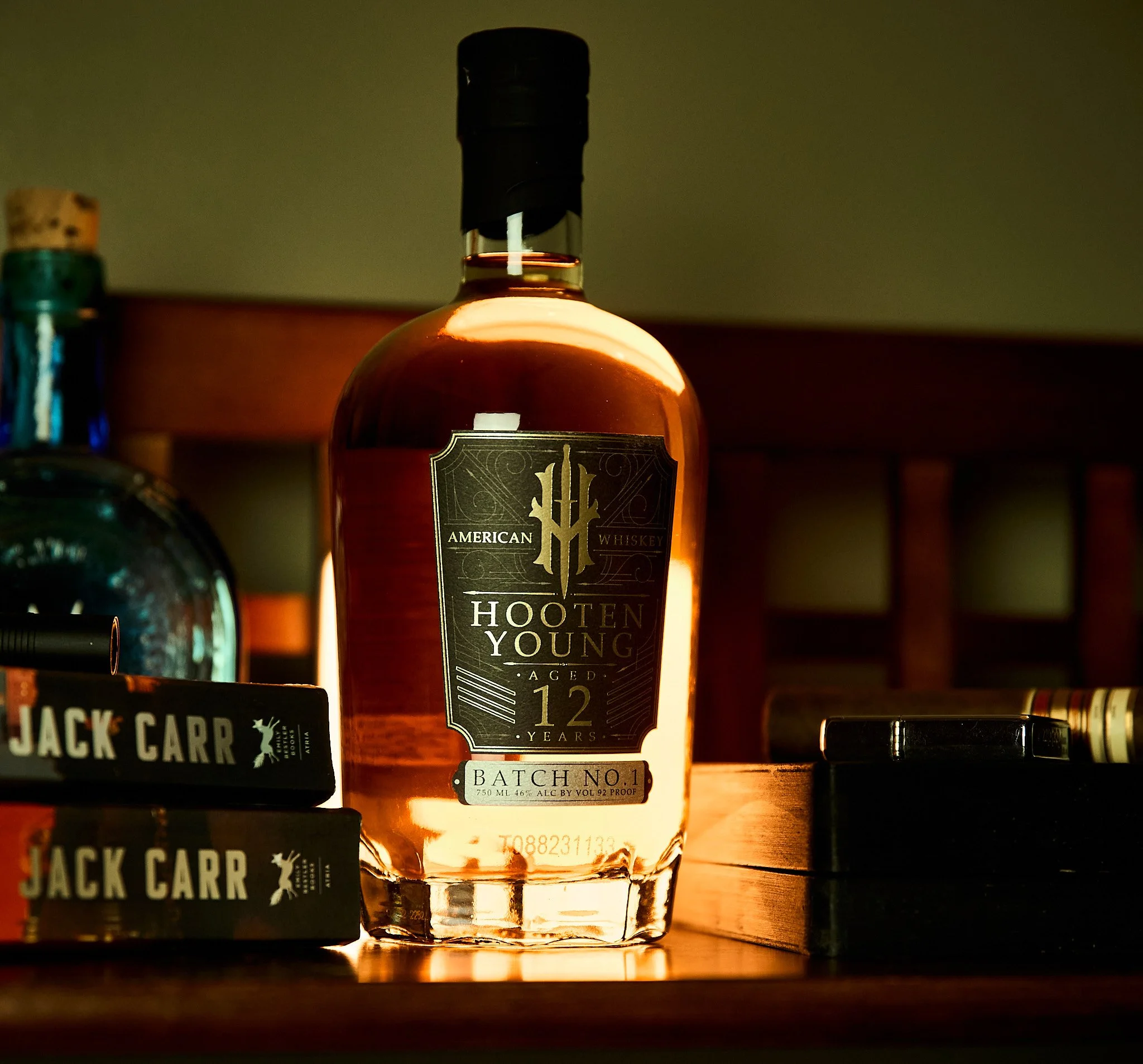 A bottle of American Hooten Young aged 12-year whiskey on a wooden surface, with Jack Carr books stacked beside it and other objects partially visible around.