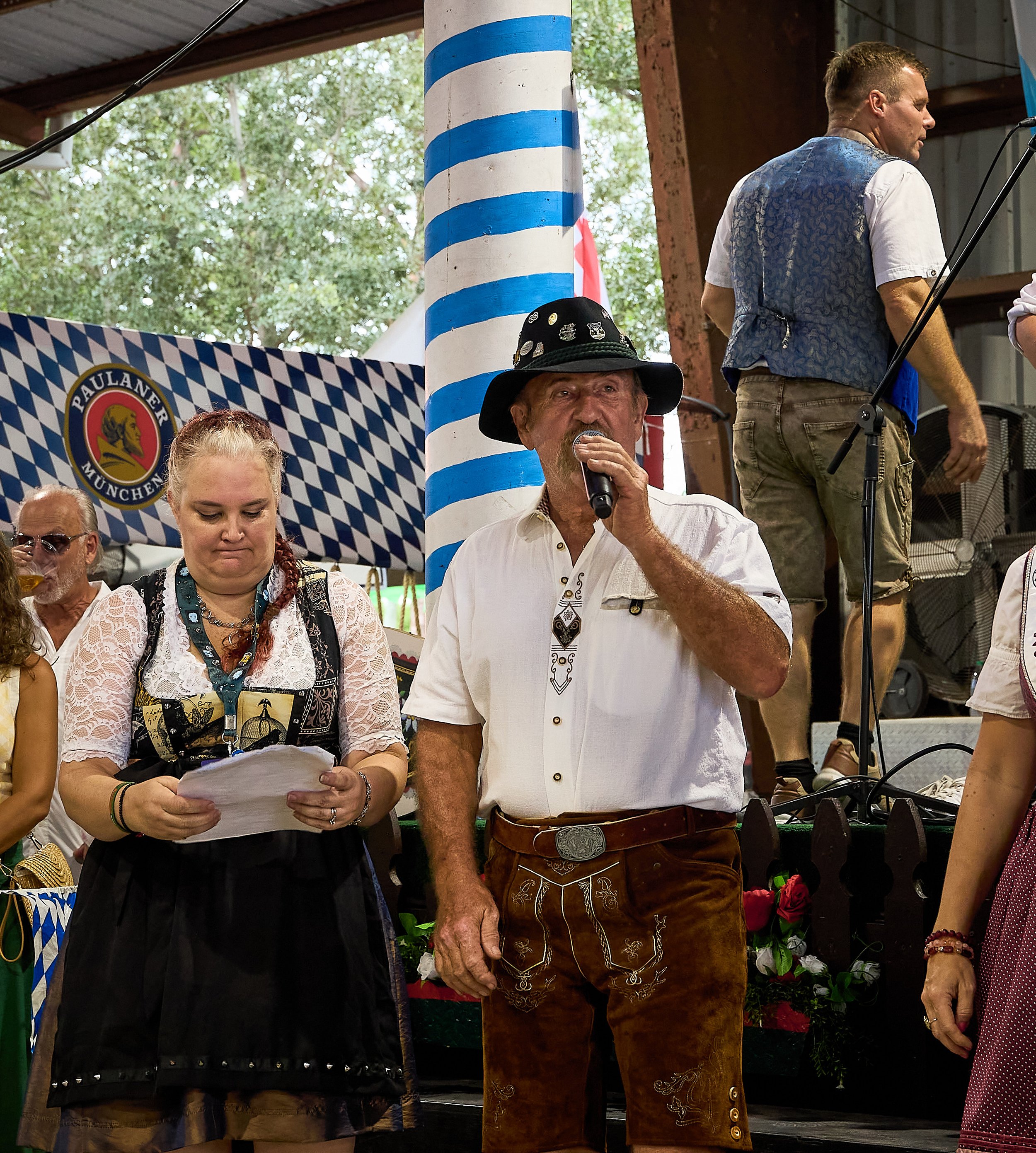 A group of people at a German cultural event, with one man speaking into a microphone and others dressed in traditional Bavarian attire, standing under a tent with a blue and white checkered banner and a striped pole in the background.