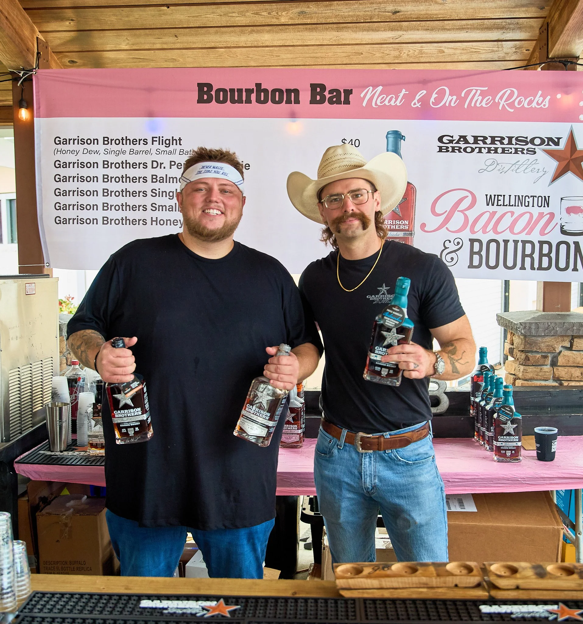 Two men standing behind a bar holding bottles of whiskey, smiling for the photo, under a banner advertising Garrison Brothers bourbon at an event.
