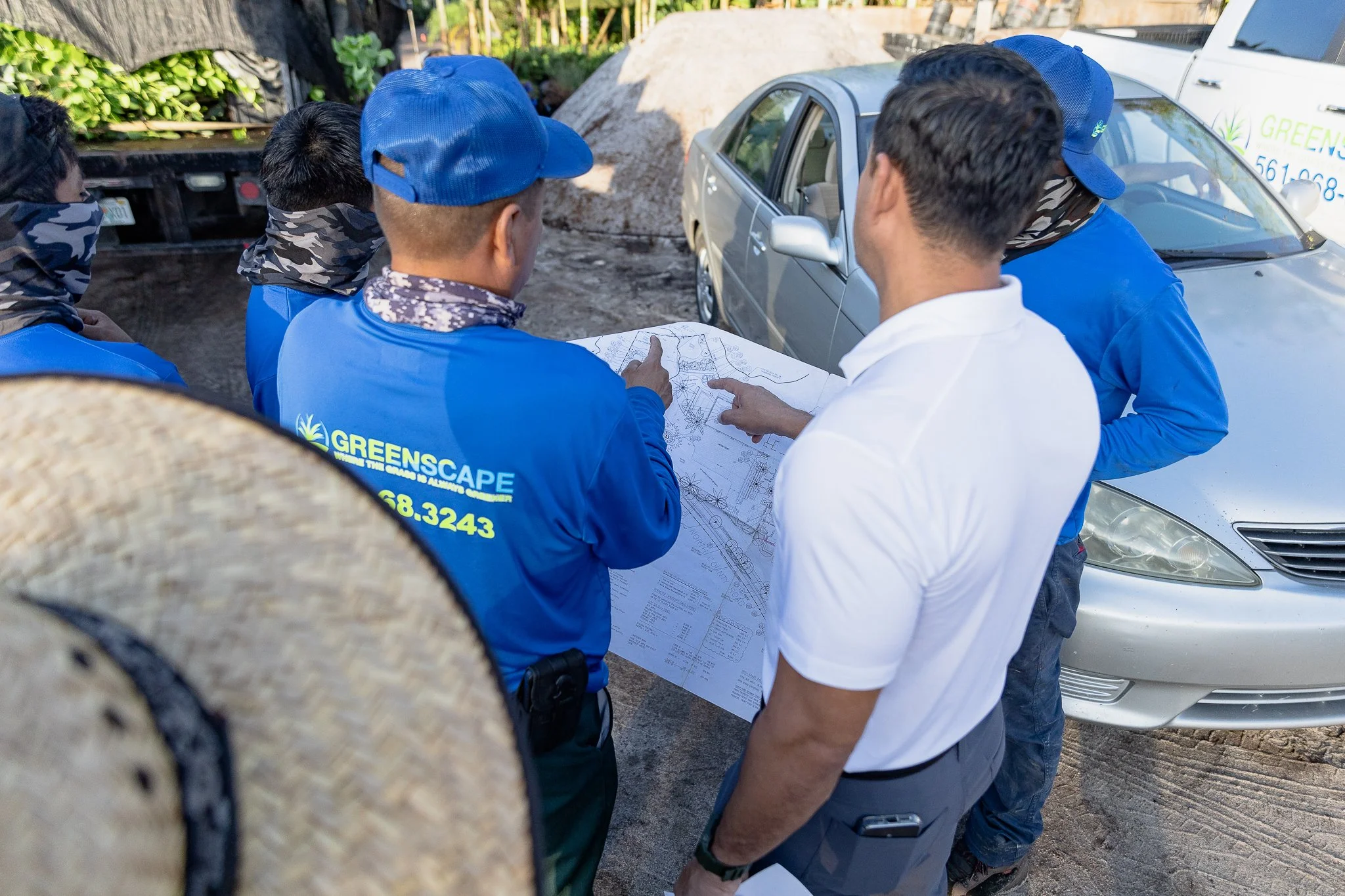 Group of construction or landscaping workers and a supervisor gathered outdoors around a large blueprint, discussing plans. Several individuals are wearing blue shirts with 'GREENS CAPE' on the back, some with camouflage neck gaiters, while one perso