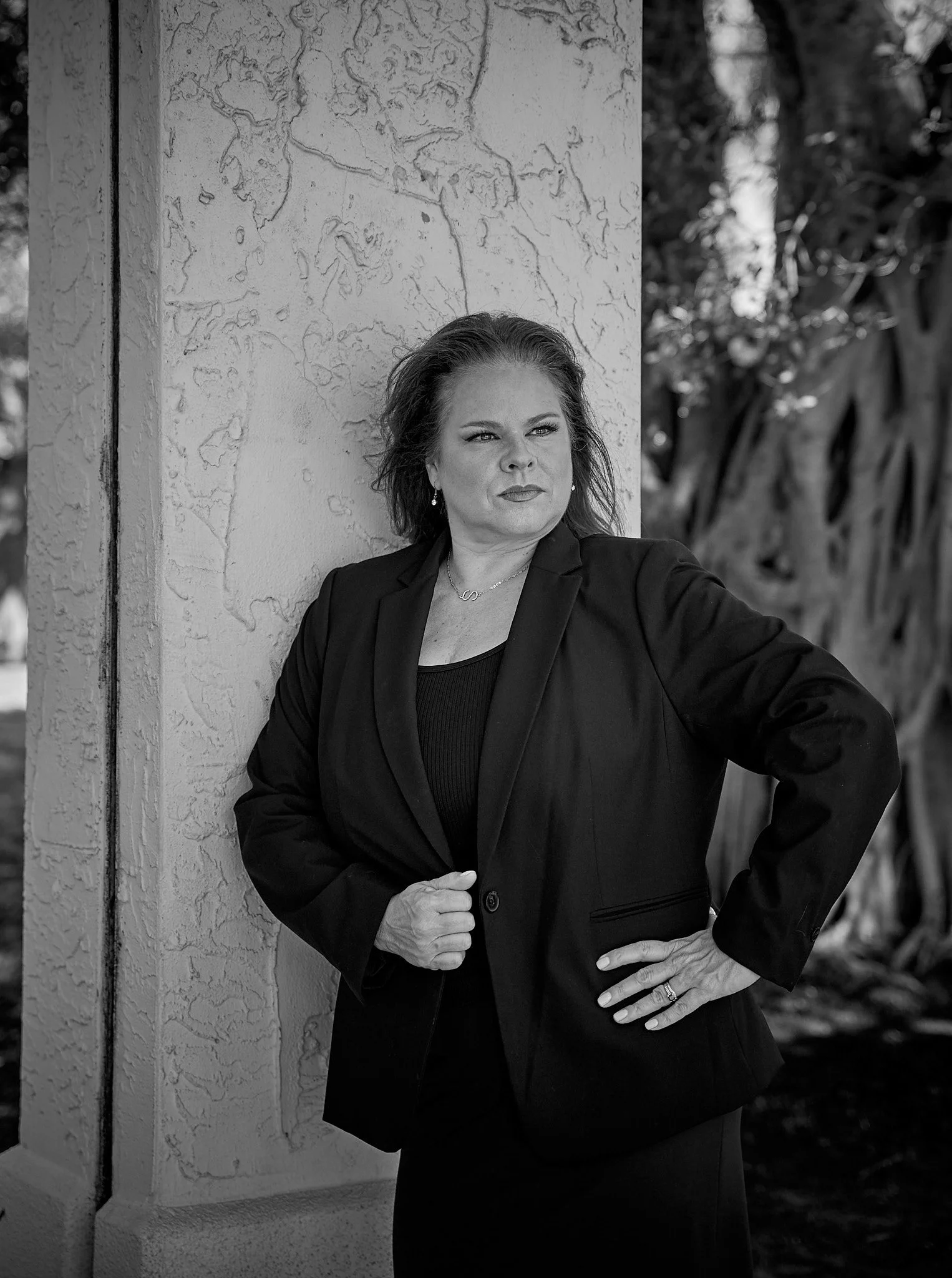 A woman in a black blazer and top stands outdoors, leaning against a textured wall with a world map design, with trees in the background.