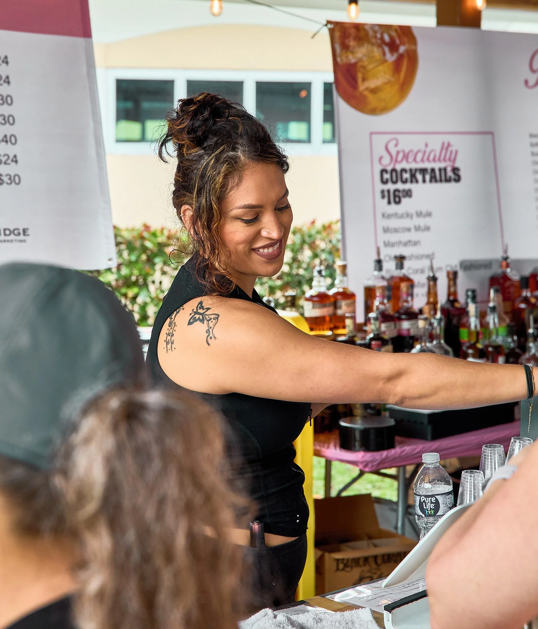 A smiling female bartender serving drinks at an outdoor booth with a colorful menu and bottles in the background.