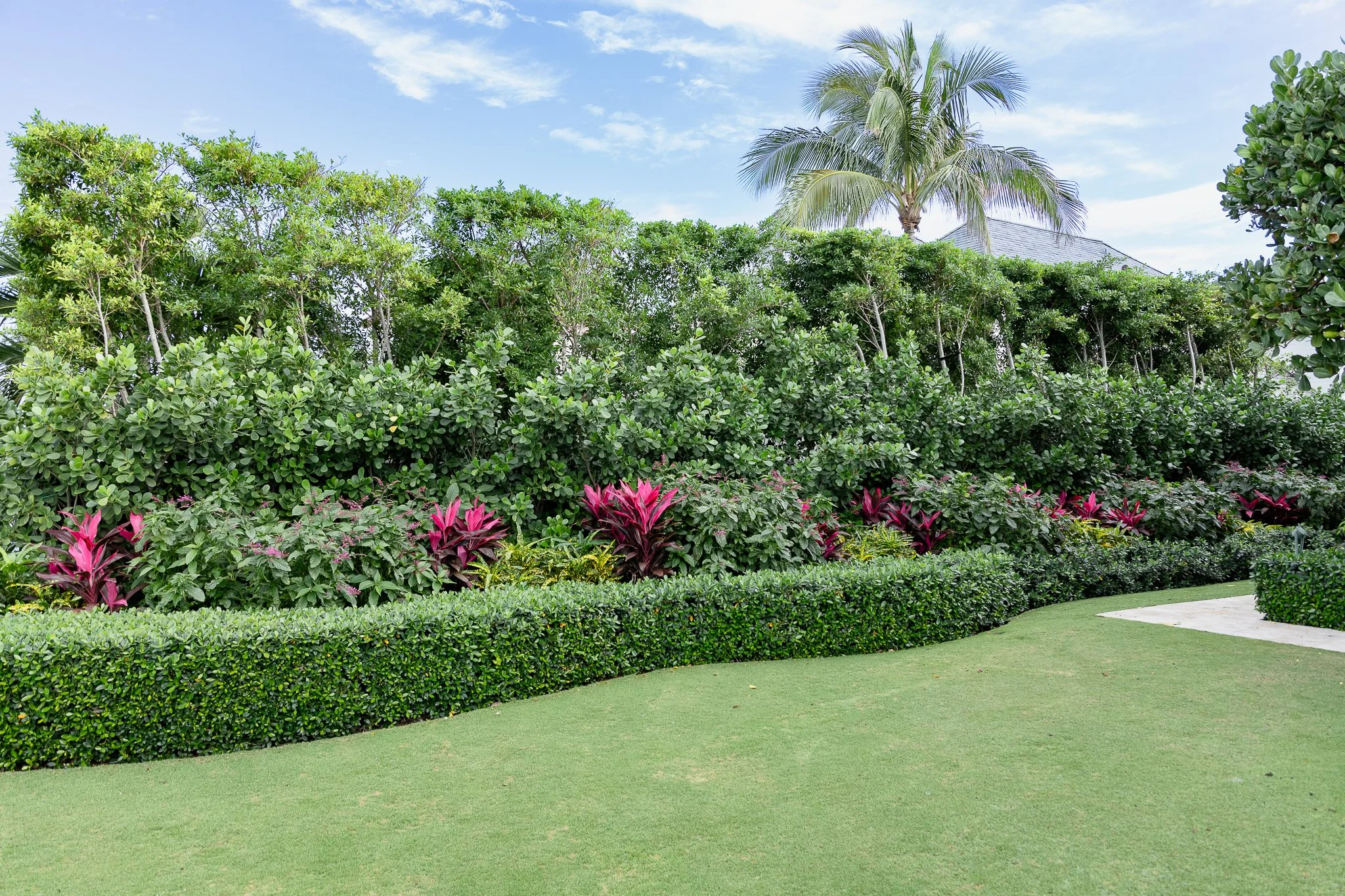 A well-maintained garden with lush green bushes, pink plants, and a curved concrete pathway, with taller trees and a house with a gray roof in the background under a partly cloudy sky.