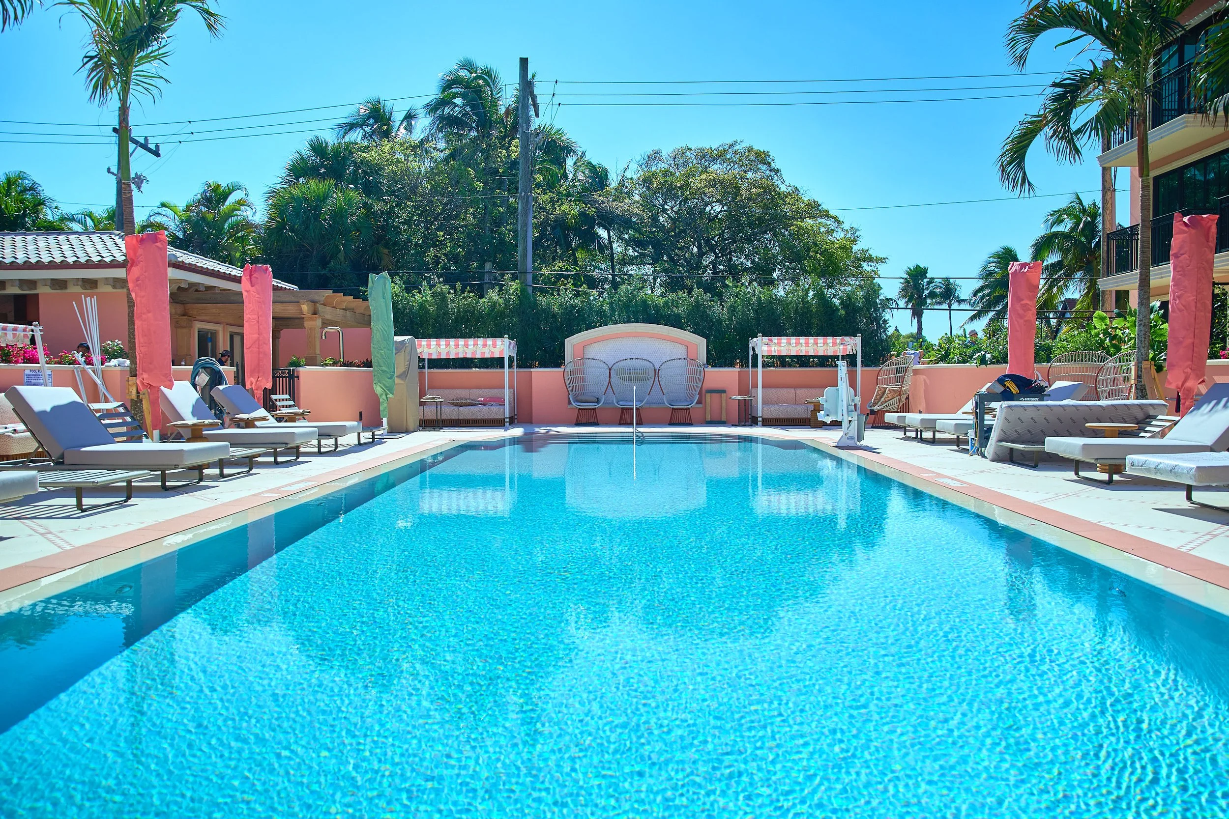 A sunny outdoor swimming pool with clear blue water, surrounded by white lounge chairs, colorful umbrellas, and tropical trees in the background.