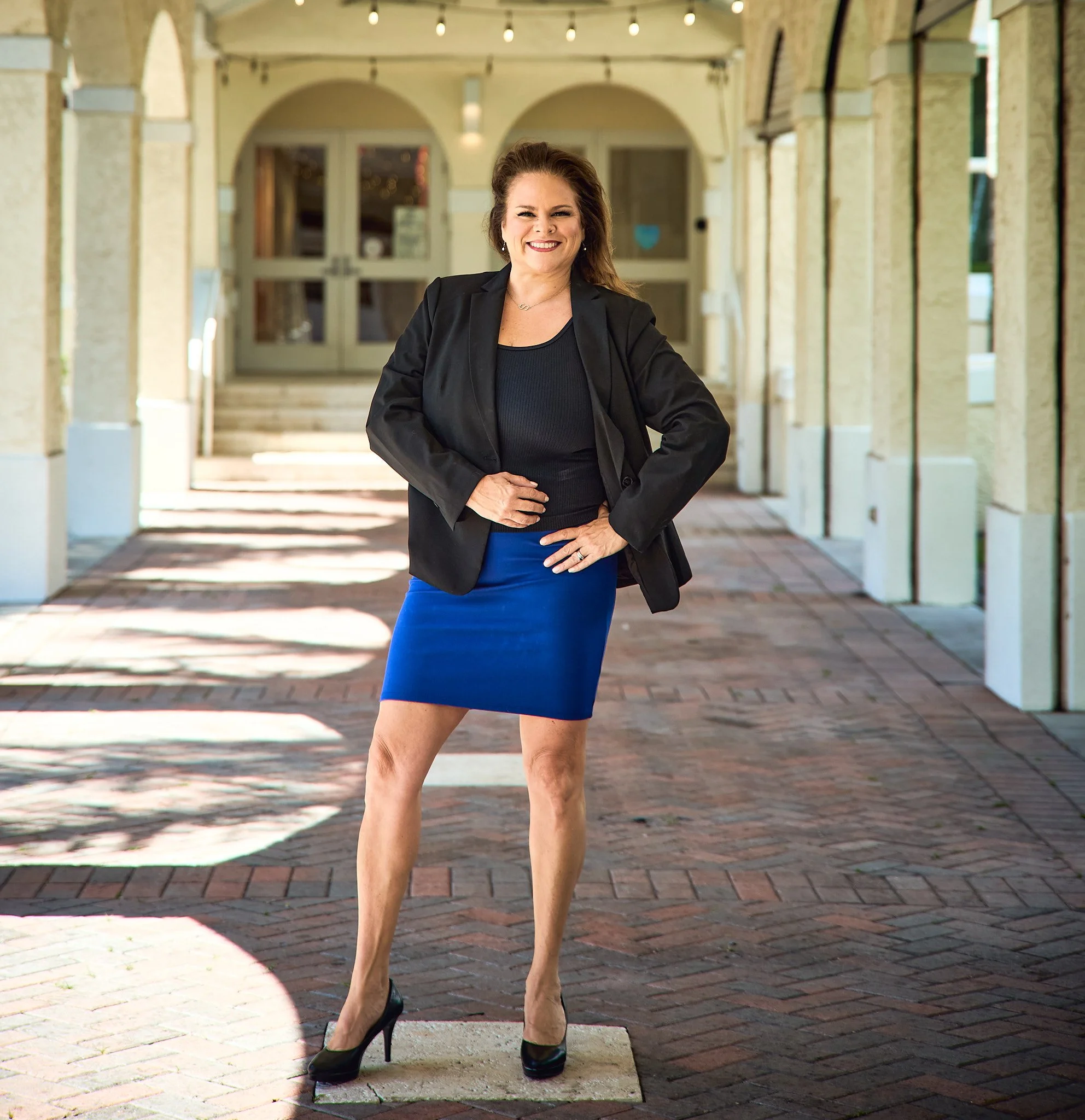 A woman in black blazer and skirt standing outdoors on a brick-paved walkway, smiling with hands on hips, against a background of arched doorways and columns.