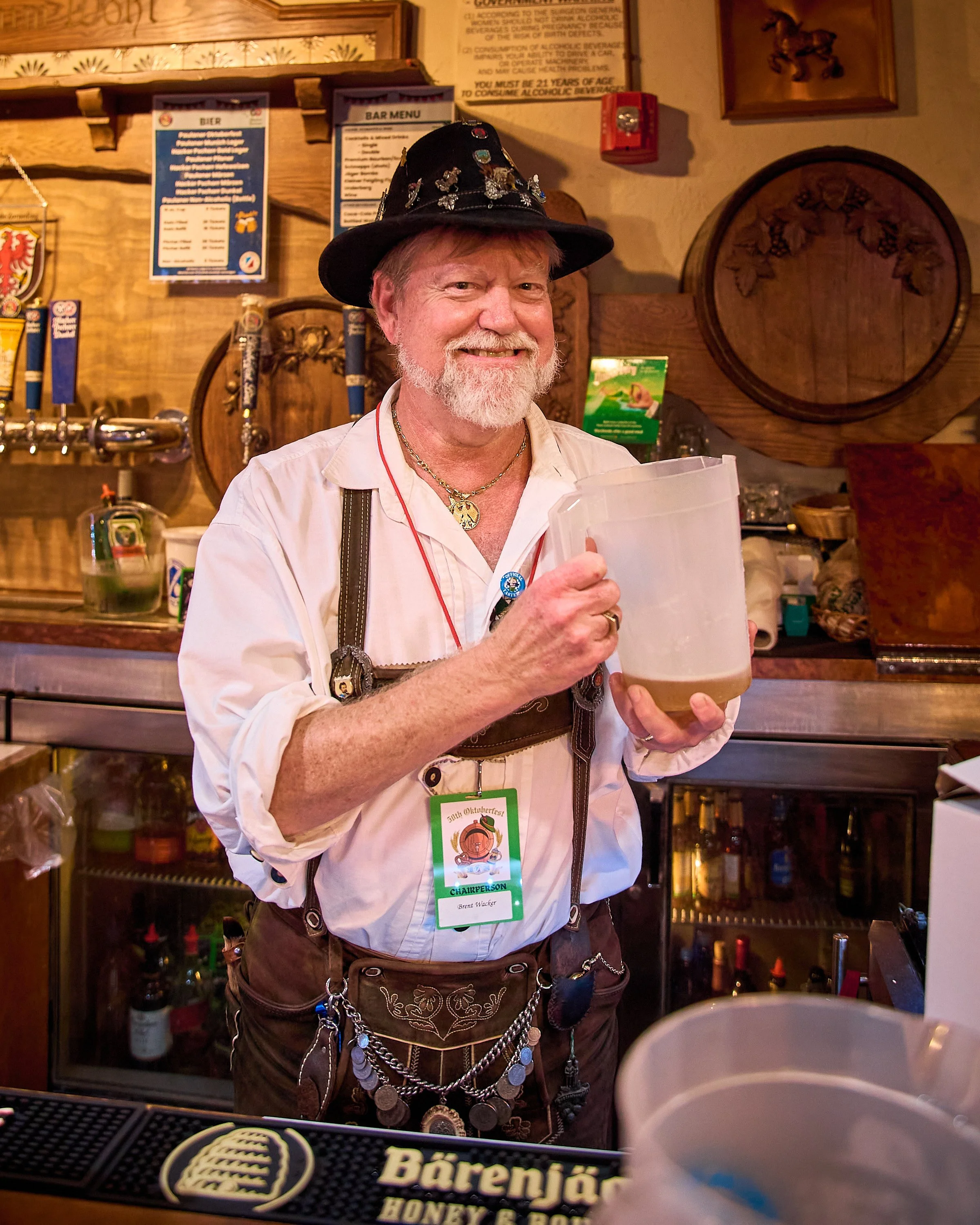 A smiling man dressed in traditional Bavarian attire, holding a pitcher of beer, standing behind a bar in a pub. He is wearing lederhosen, a white shirt, suspenders, a decorative hat with pins, and a necklace, with beer taps and signs in the backgrou