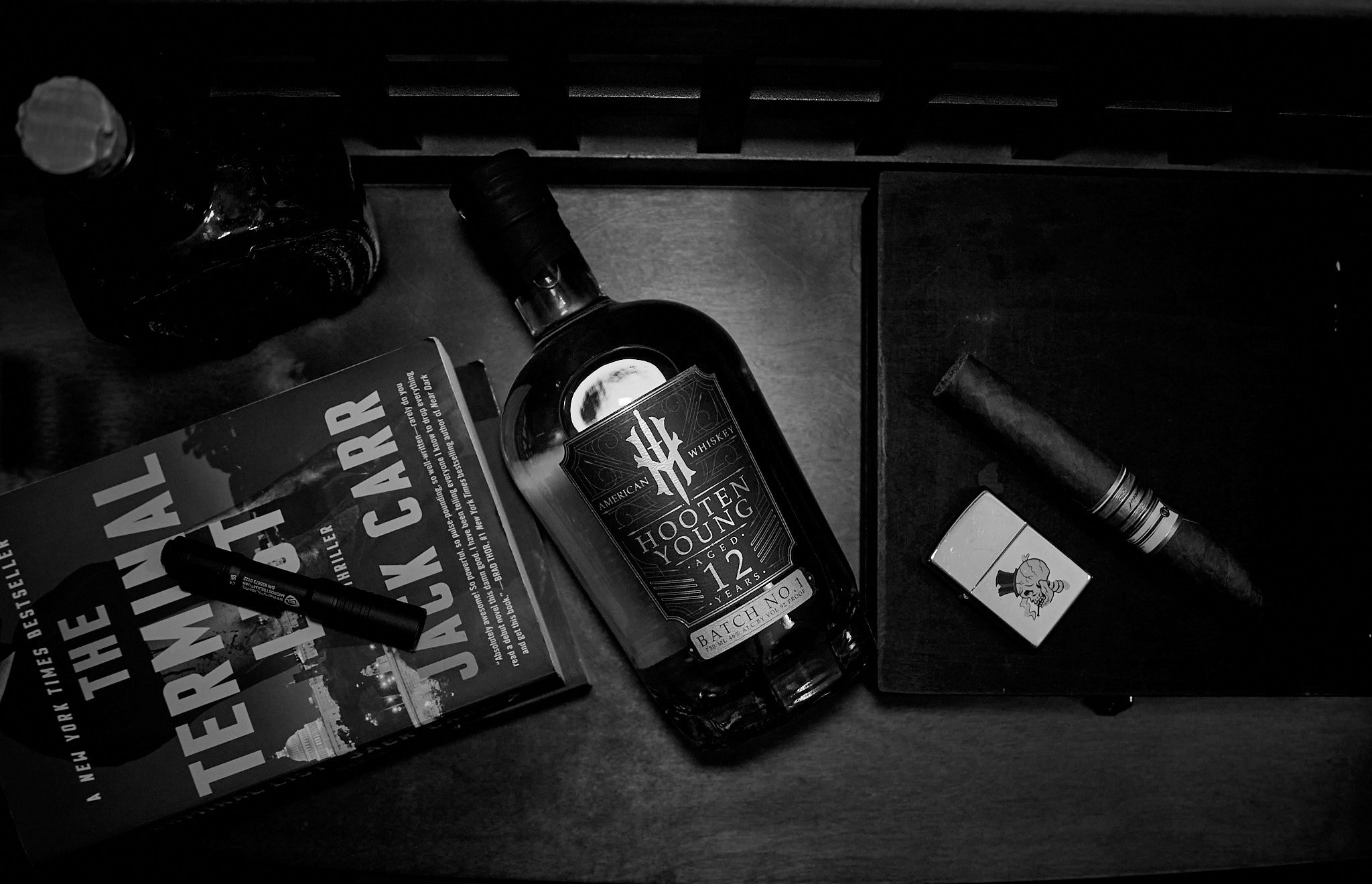Black and white photo of a wooden table with a book titled 'The New York Times Best Seller', a marker, a bottle of American whiskey labeled 'Hooting Yount, aged 12 years', a cigar, a lighter, and a cigar tube on it.