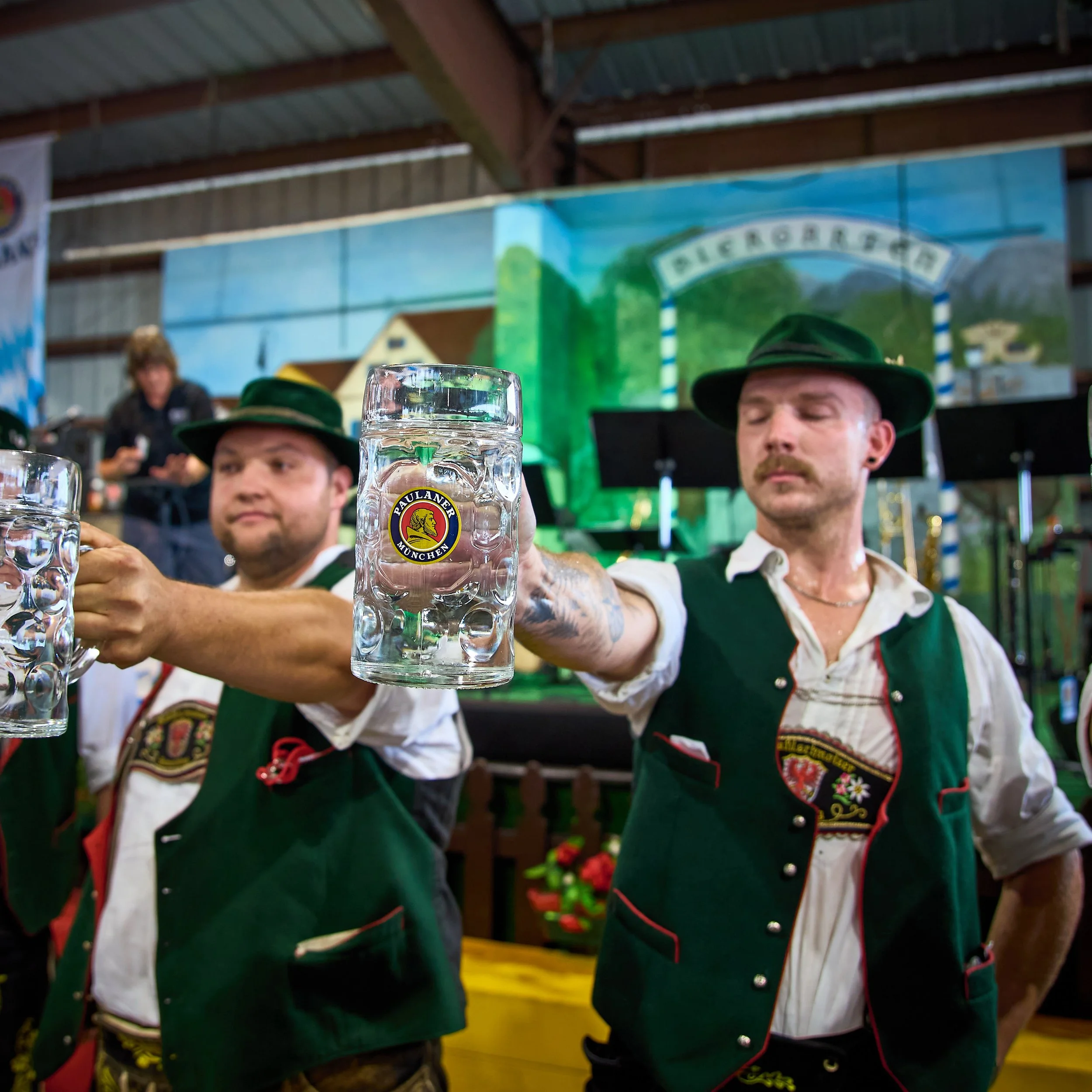 Two men wearing traditional Bavarian outfits, including green hats and vests, holding up large steins of beer during a celebration or festival.