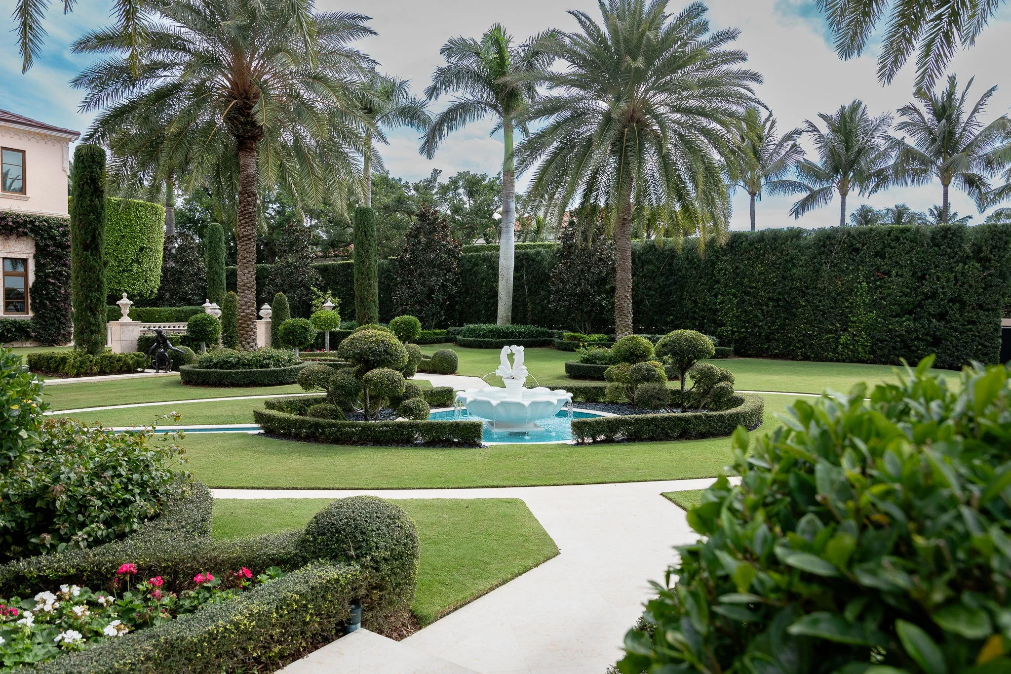 A landscaped garden with a central fountain, surrounded by neatly trimmed bushes, tall palm trees, and a large hedge in the background.