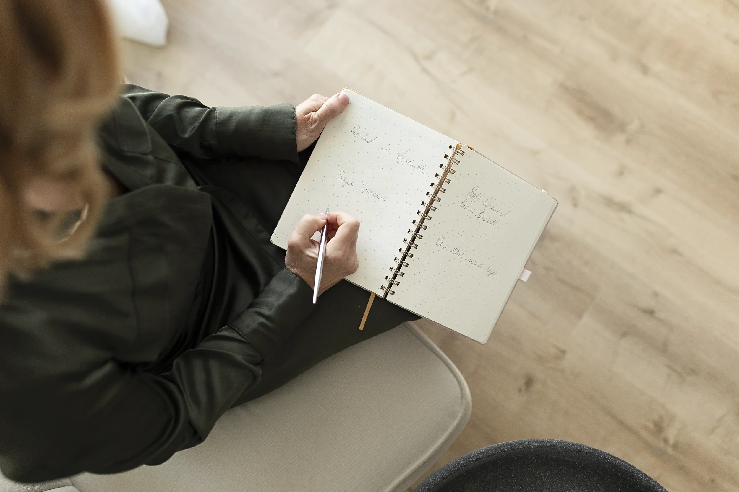 A person with blonde hair, wearing a black jacket, sitting on a white chair, writing in a spiral notebook. The notebook contains handwritten notes, and the person's hand is holding a pen. The setting has a wooden floor.
