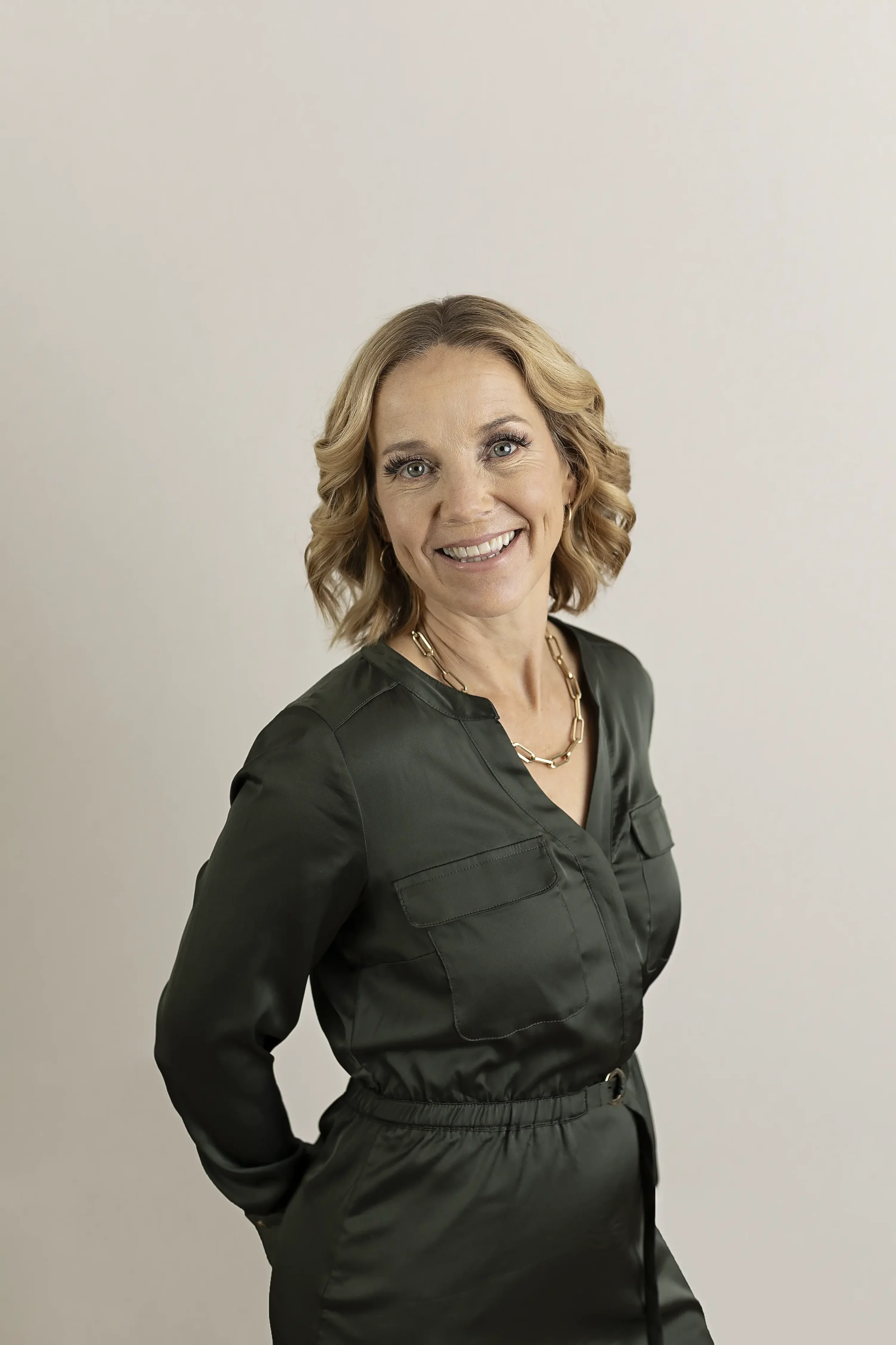 A woman with shoulder-length wavy blonde hair, smiling, wearing a black jumpsuit with a gold chain necklace and standing against a plain light-colored background.