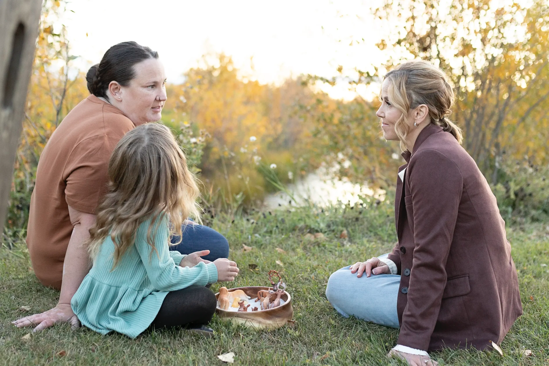 Three women and a young girl sharing a moment outdoors on a grassy area near a body of water, with trees showing autumn colors in the background. One woman with dark hair in a bun is sitting and leaning forward, another woman with blonde hair in loose waves and wearing a maroon jacket is sitting on the grass, and a young girl with long blonde hair is sitting between them, playing with small figurines.