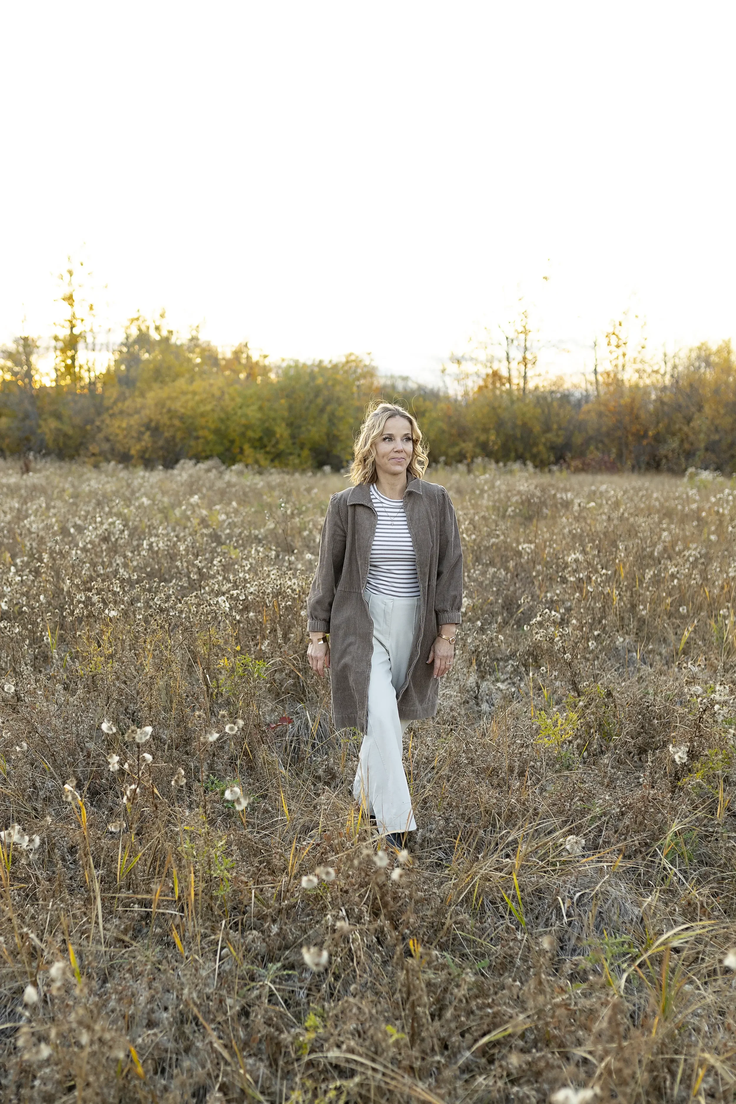 A woman walking through a field of dry grass and wildflowers at sunset, wearing a brown coat, striped shirt, and white pants.
