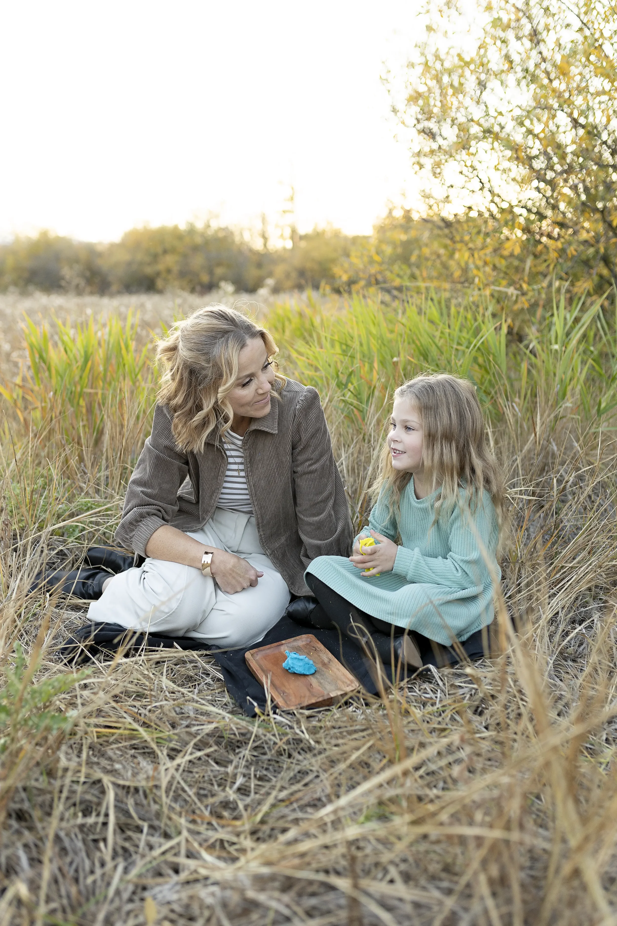 A woman and a young girl sitting on a black mat in a grassy outdoor area with tall grass and trees, during what appears to be late afternoon or early evening.