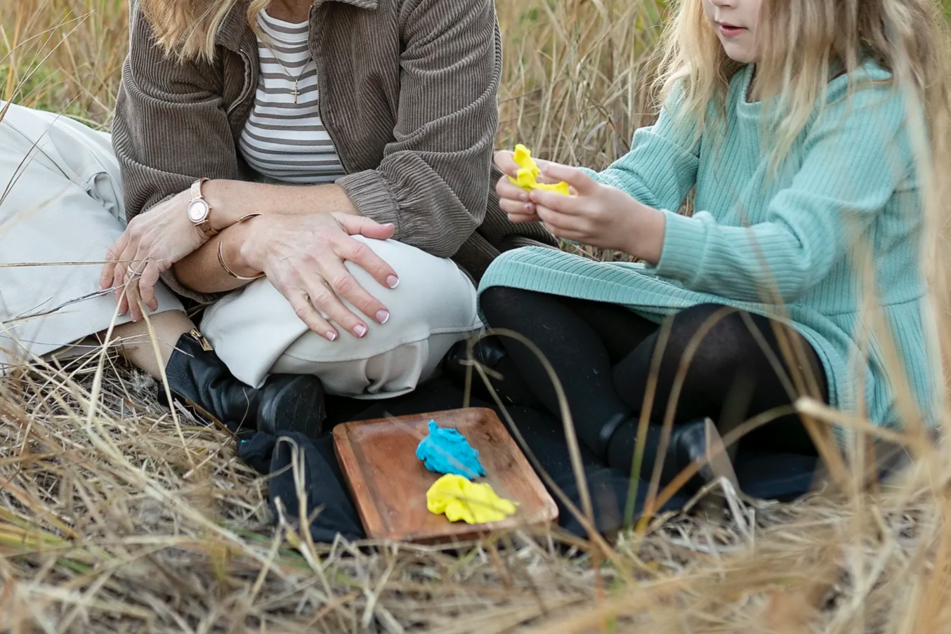 A woman and a girl sitting in a field, engaged in finger-painting activity with yellow and blue paint on a small wooden board a