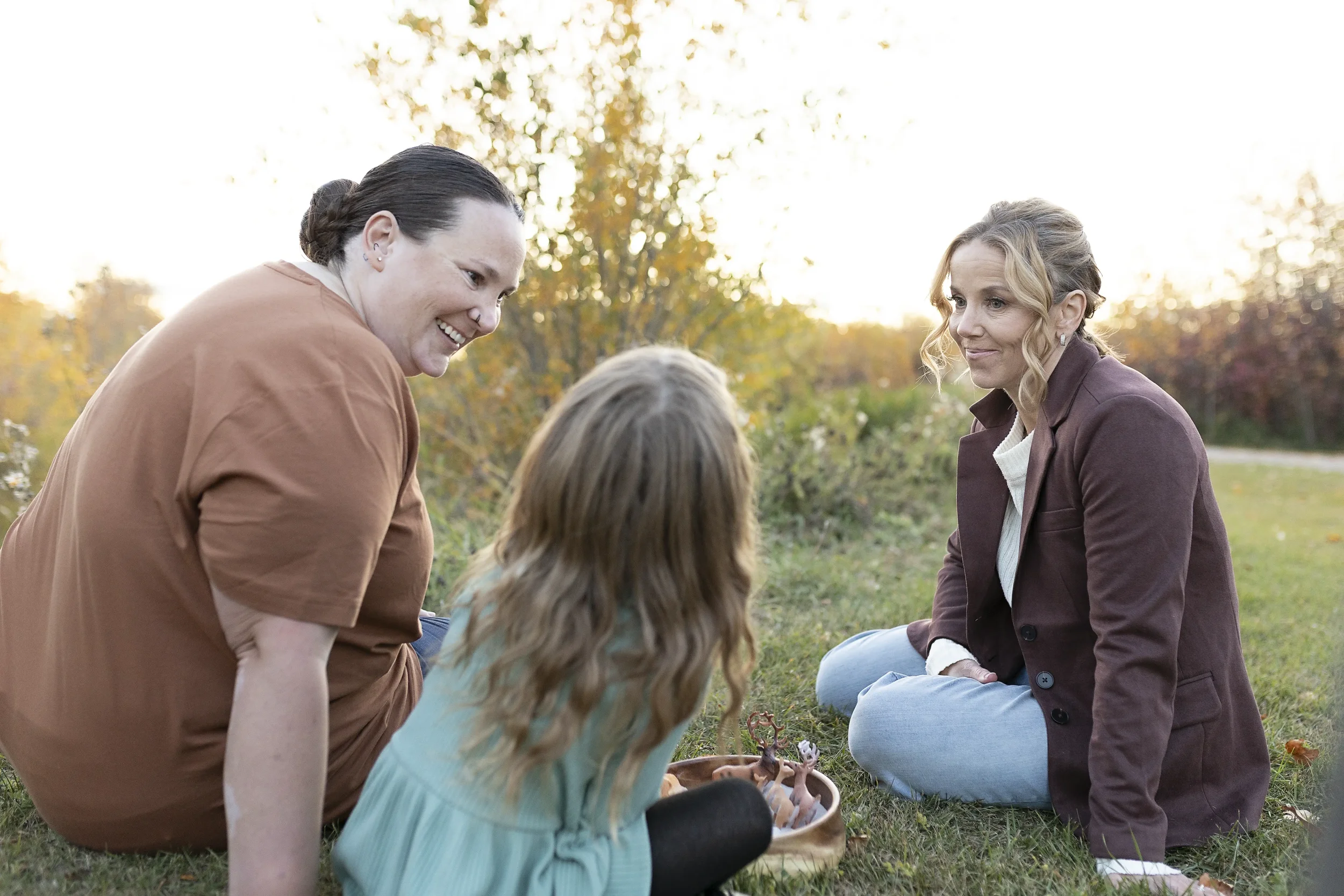 Three women sitting and kneeling on grass in a park during autumn, engaging with a young girl with long blonde hair as she holds a small bowl with toy dinosaurs.