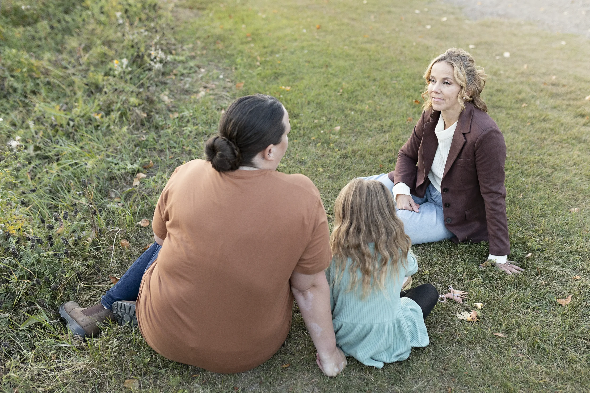 Four people, two adults and two children, sit on grass in a park-like setting, engaged in a conversation.