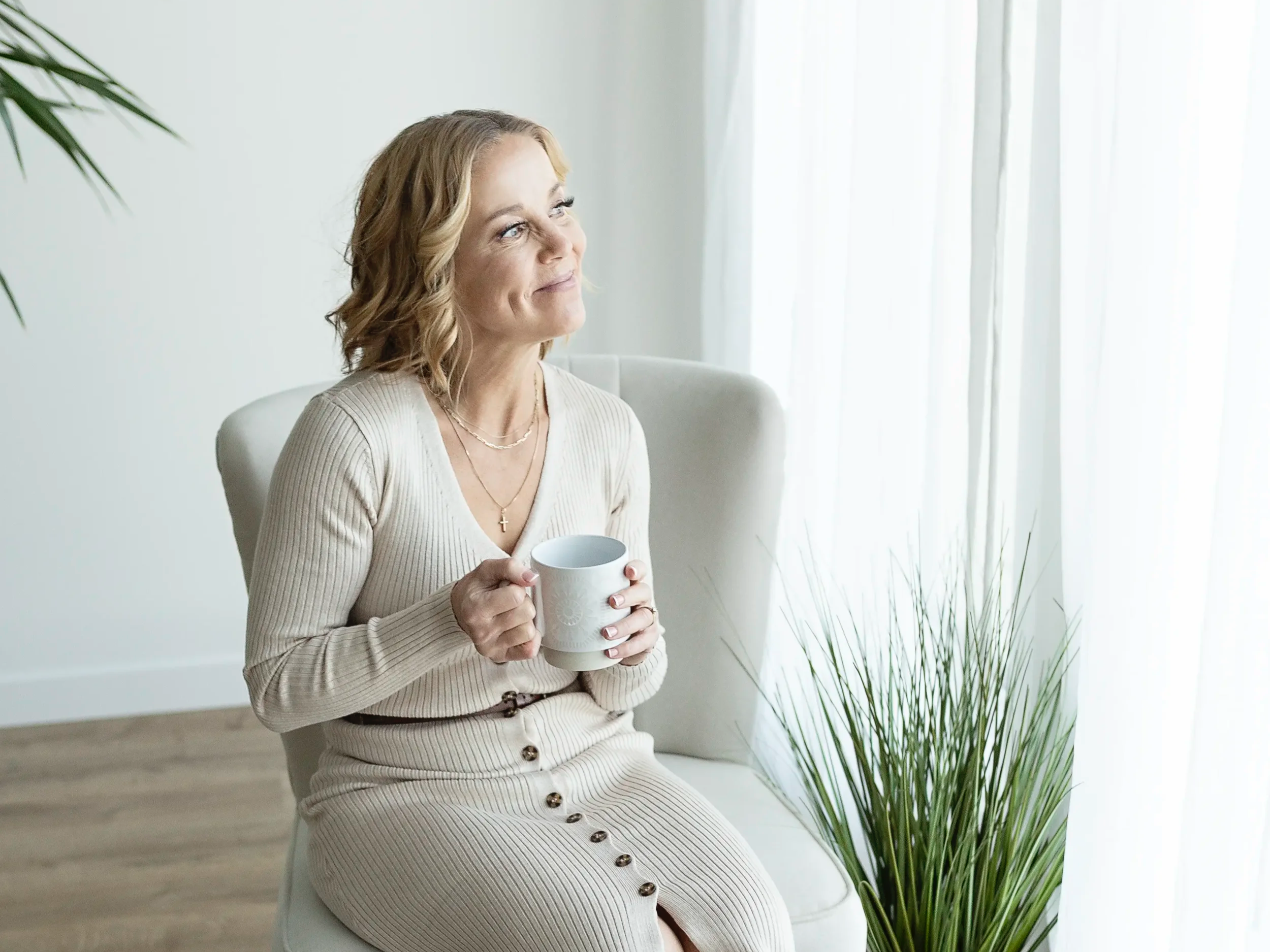 A woman with short, wavy blonde hair sitting in a white armchair, holding a white mug and gazing out a window with white curtains, along with a green potted plant nearby.