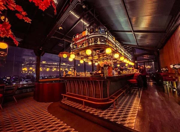Interiores de un bar elegante con iluminación cálida, bar bordeado de bancos de madera, techo negro, grandes ventanas con vista a la ciudad y decoración moderna con flores de colores.
