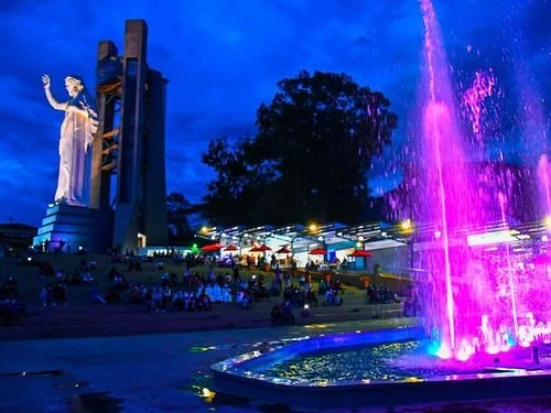 Escenario con estatua de una figura femenina, fuente de agua iluminada con luces coloridas, y grupos de personas disfrutando en el parque por la noche.