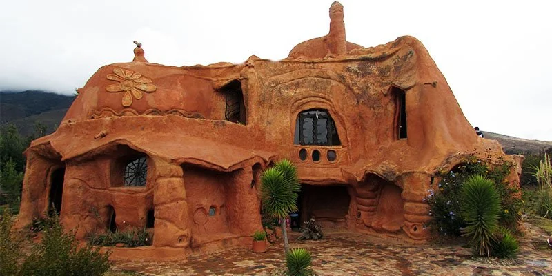 Casa estilo adobe de colores terracota con detalles decorativos en forma de flor, rodeada de plantas y en un entorno montañoso.