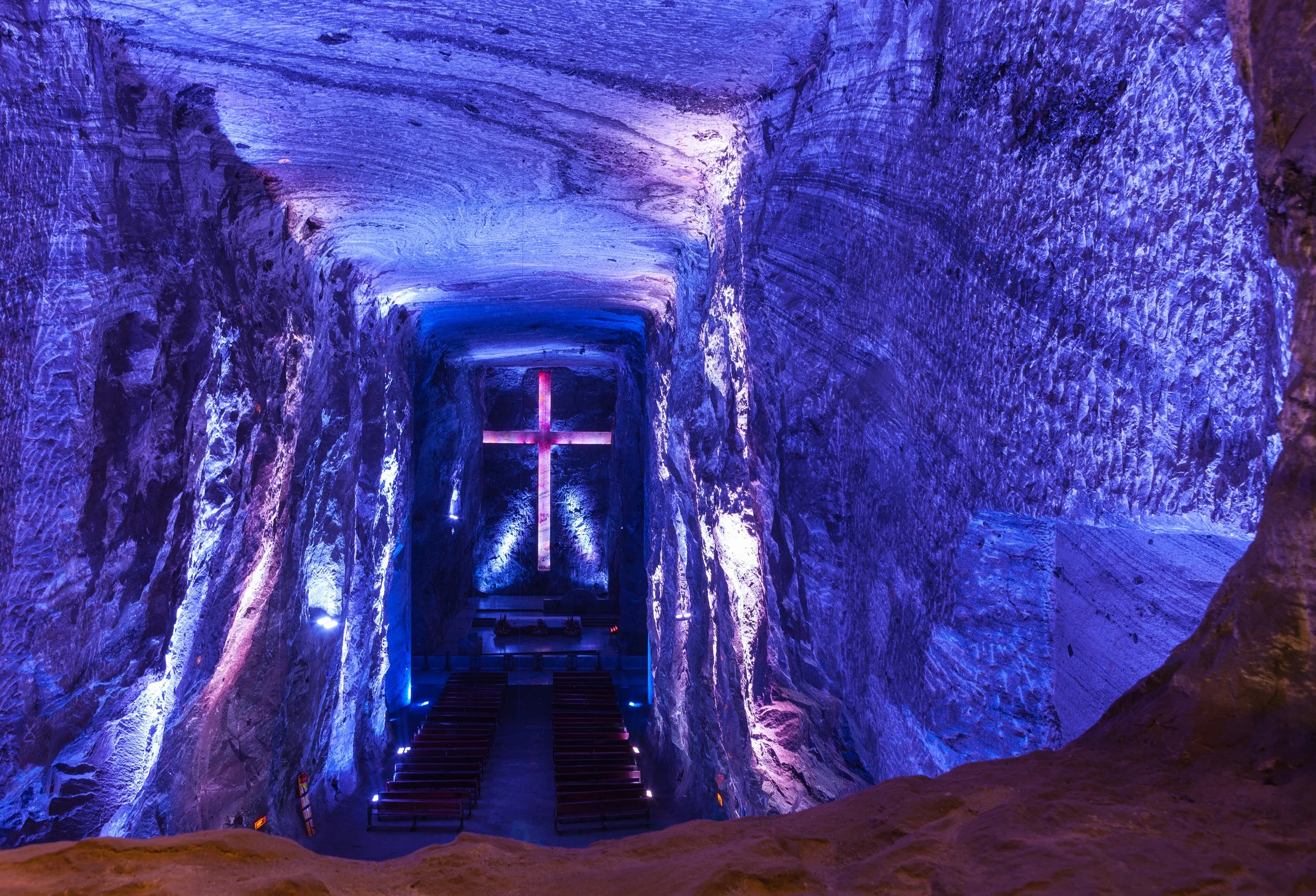 Interior de una cueva con paredes iluminadas en tonos morados y azules, y una cruz luminosa en el fondo.