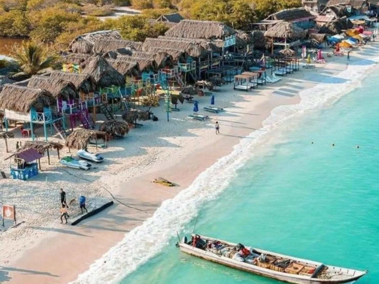 Vista aérea de una playa con cabañas de palapa, sombrillas de paja, palmeras, y un barco en el agua. Hay algunas personas caminando en la arena y en el agua.