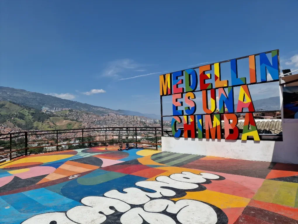 Vista de un mirador en Medellín con un cartel colorido que dice "Medellín es una chimba", en un espacio decorado con pintura vibrante en el suelo y un fondo de montañas y ciudad bajo un cielo despejado.