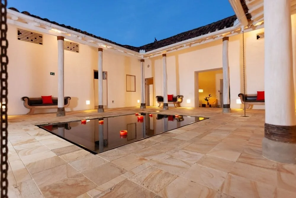 Patio interior con fuente reflejante, columnas blancas, sillas de madera con cojines rojos, y decoraciones en la noche.