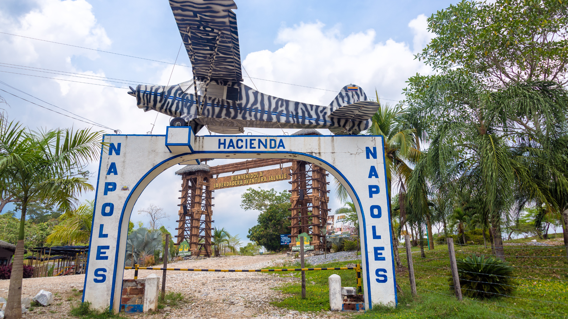 Entrada a una hacienda llamada Nápoles con un arco pintado de blanco y azul, un avión decorativo en la parte superior, y estructuras de madera. Hay palmeras y vegetación tropical alrededor.