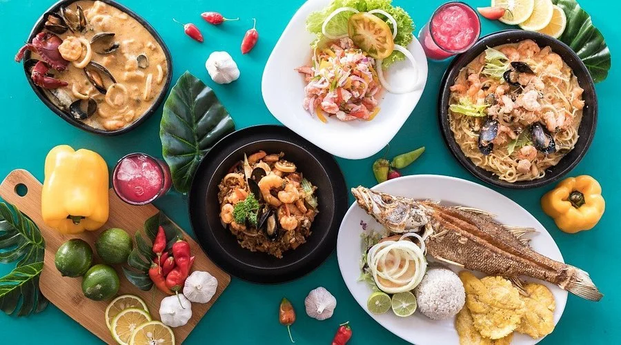 Plato de pescado con limón, arroz y ensalada, junto a pasta con mariscos, sopa cremosa con mariscos y verduras, y una ensalada con lechuga, tomate y cebolla. Decoración con verduras, limones y chiles.