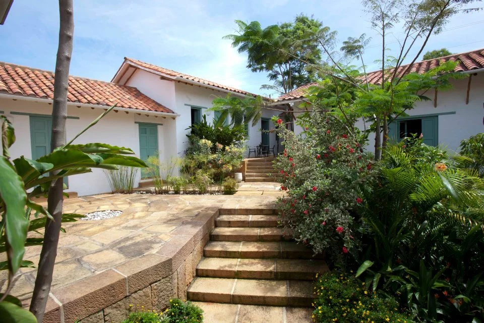 Casa colonial con techo de tejas rojas, escaleras de piedra, jardín con plantas verdes y flores, y cielo despejado.