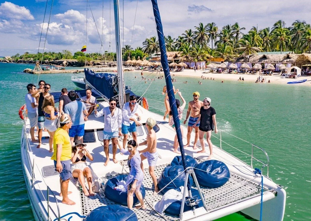 Grupo de personas disfrutando en un yate en una playa con palmeras, sombrillas y bañistas en el agua.