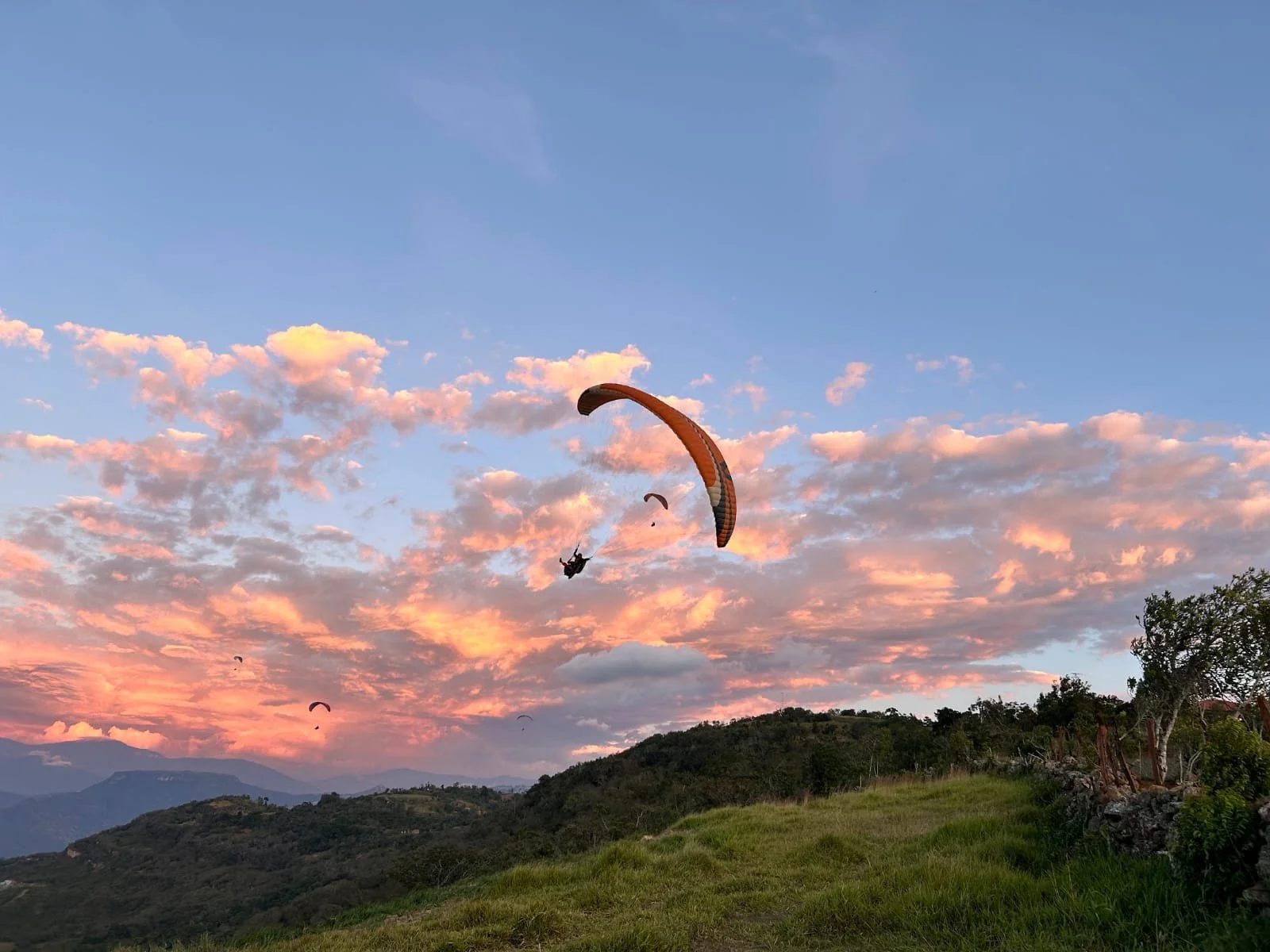 Parapentes volando sobre colinas verdes bajo un cielo con nubes rosadas y azules al atardecer.