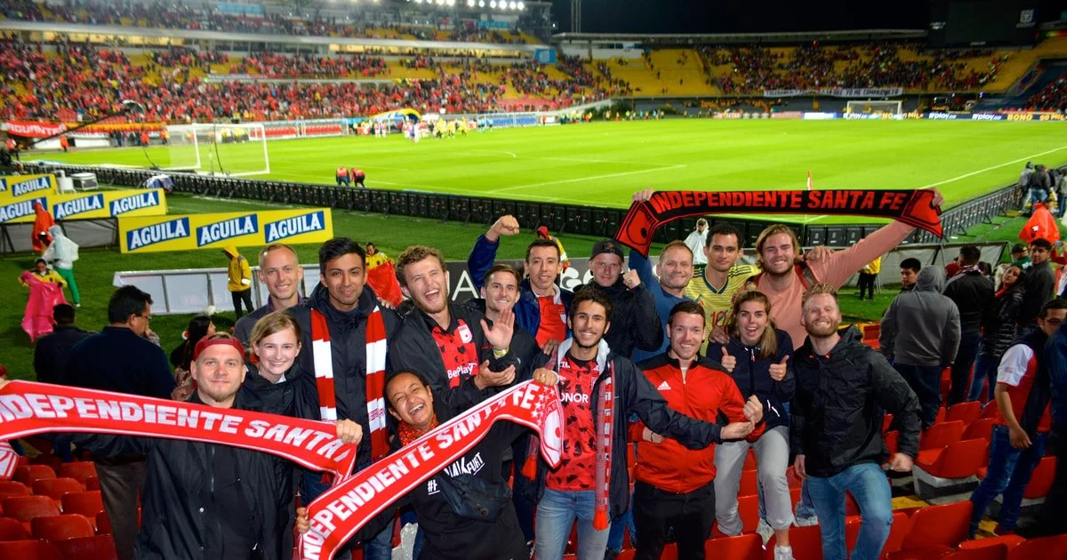 Grupo de personas en un estadio de fútbol, celebrando con bufandas del equipo Independiente Santa Fe, en un partido.