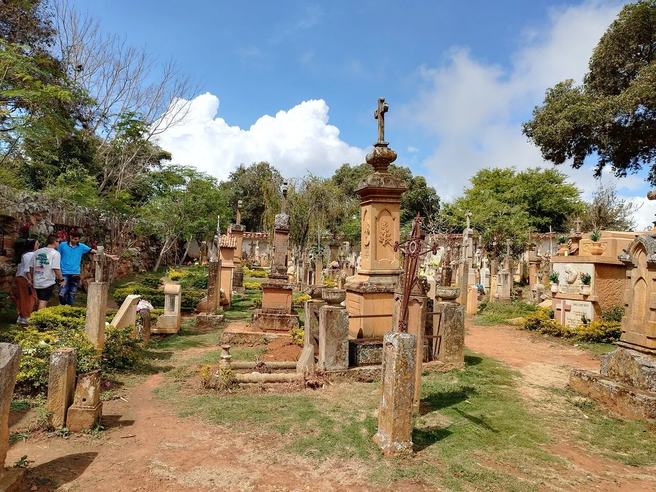 Grupo de personas paseando por un cementerio con tumbas y cruces, rodeado de árboles y césped, bajo un cielo parcialmente nublado.