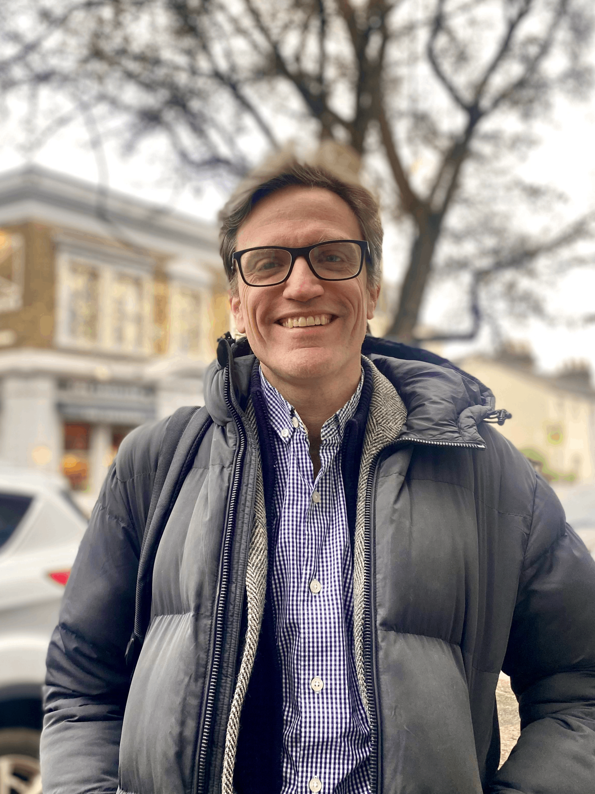 A man wearing glasses, a black puffy jacket, and a blue checkered shirt standing outdoors with a city street in the background, trees, and cars.