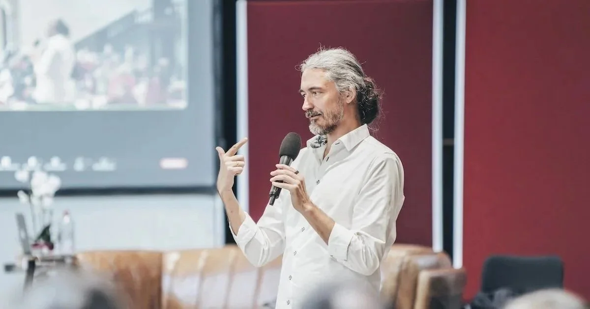 A man with gray hair and a beard, wearing a white shirt, speaking into a microphone during a presentation or lecture in a conference room.