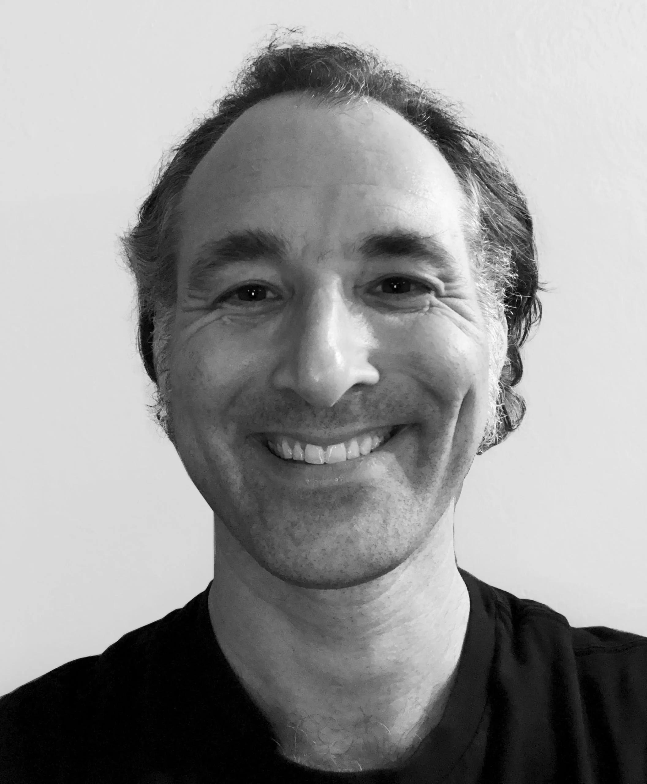 Close-up black-and-white portrait of a smiling man with short, curly hair and a beard, wearing a dark shirt against a plain background.