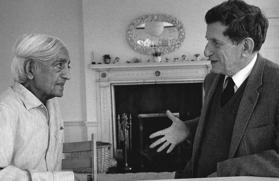 A black and white photo of two men having a conversation in a living room, with a fireplace and a decorative mirror on the wall behind them. One man has white hair and is wearing a light-colored shirt, while the other man has dark hair and is dressed in a dark blazer with a sweater and tie.