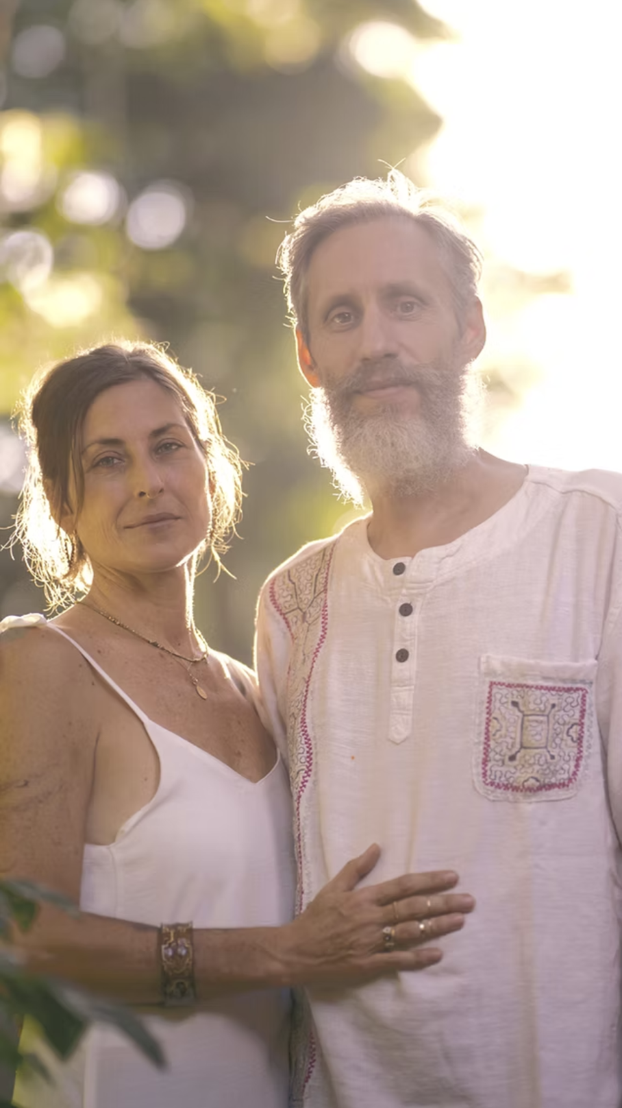 A man and woman standing closely outdoors during sunset, with warm sunlight behind them. The woman has brown hair and is wearing a white sleeveless dress and jewelry. The man has gray hair and a beard, wearing a light-colored shirt with embroidery details.