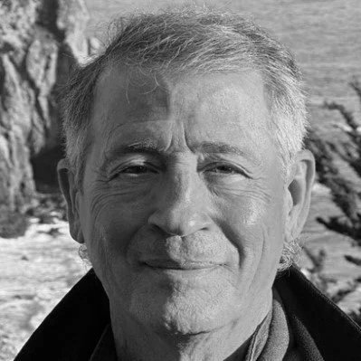 A black and white photo of an older man with short, gray hair smiling outdoors, with trees and water in the background.