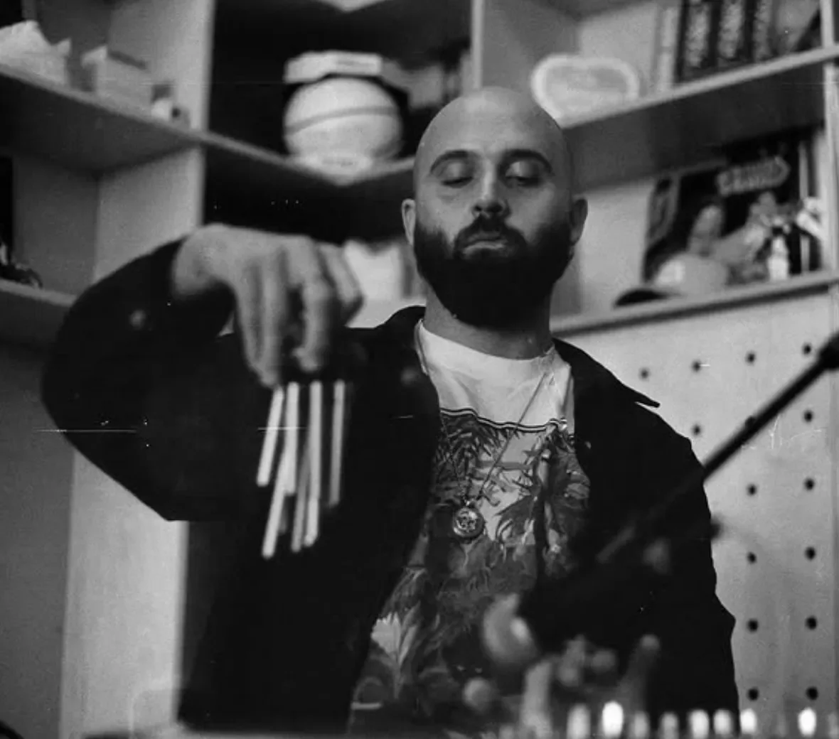 A man with a beard and shaved head looking down with eyes closed, holding a set of multiple knuckle-duster brass knuckles in a workspace or garage with shelves and boxes in the background.