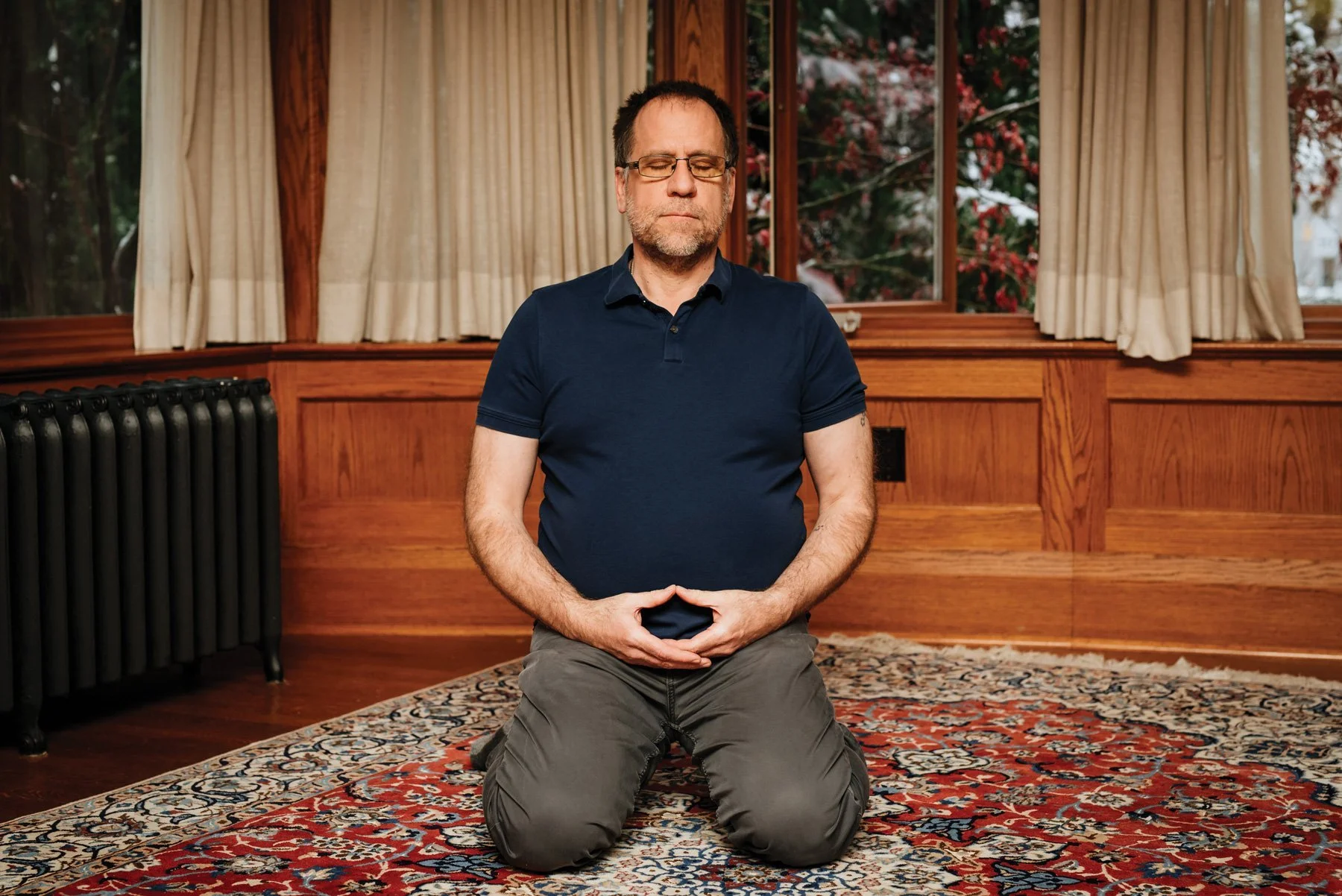 A man kneeling on a patterned rug, meditating with eyes closed and hands forming a mudra, in a cozy wooden room with large windows and beige curtains.