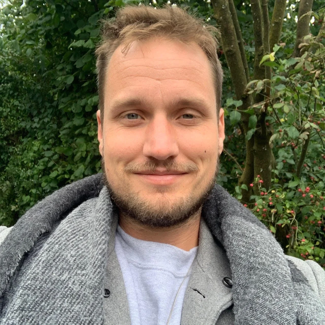 A smiling man with short hair and a beard, wearing a gray coat and a light-colored shirt, standing outdoors in front of green trees.